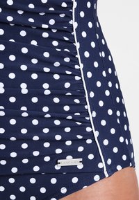Maillot de bain bleu marine à pois, avec des pois blancs, un tissu texturé, des fronces sur les côtés et une étiquette métallique de la marque.