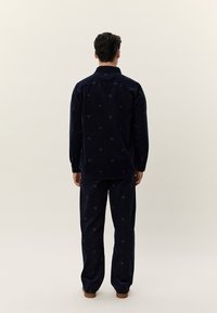 Mann stående med ryggen til, iført mørkeblått mønstret skjorte med lange ermer og matchende bukser, sammen med brune sko, foran en ensfarget bakgrunn.