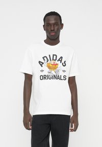 Balti medvilninė marškinėliai su ryškiu grafiniu atspaudu, kuriame yra tekstas „ADIDAS ORIGINALS“ ir animacinis personažas, derinami su tamsiomis džinsais.