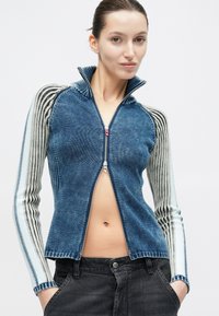 Ung kvinna klädd i blå zip-up tröja med randiga ärmar och svarta jeans, ståendes med händerna i fickorna mot vit bakgrund.