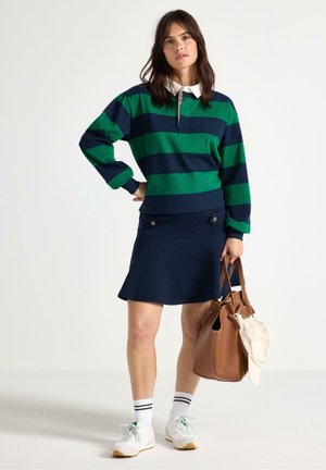 Top a maniche lunghe a righe verdi e blu con colletto bianco, abbinato a una gonna blu navy con dettagli in bottoni. Borsa marrone e sneakers.