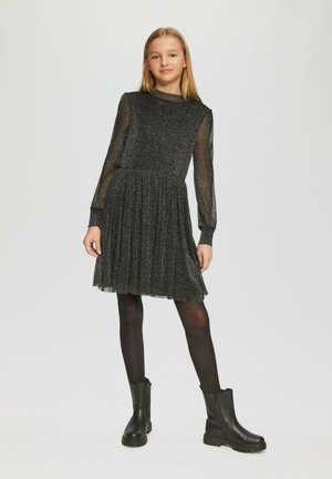 Schwarzes Glitzerkleid mit langen, durchsichtigen Ärmeln und plissiertem Rock, kombiniert mit schwarzen Knöchelstiefeln. Glattes und strukturiertes Material.