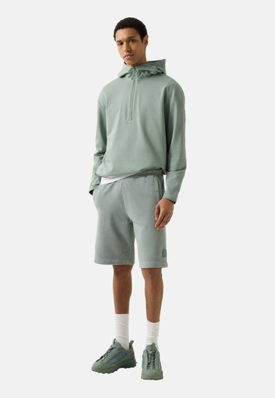 Jeune homme portant un pull à capuche vert clair, un short assorti, des chaussettes blanches et des baskets vertes, debout les mains dans les poches.