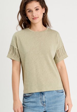 Młoda kobieta w beżowej bluzce z krótkim rękawem i koronkowymi panelami na rękawach oraz jasnych niebieskich dżinsach, stojąca na jednolitym tle.