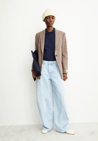 Bruine textured blazer over een navy polo, gecombineert met lichtblauwe wijde jeans en een witte gebreide beanie. Houdt een navy tas vast.