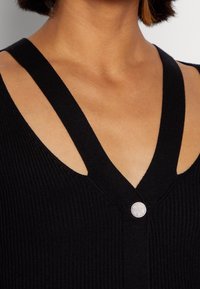 Top nero a coste con scollatura a V profonda e spalline larghe. Presenta un dettaglio circolare e luccicante a forma di bottone sul collo.