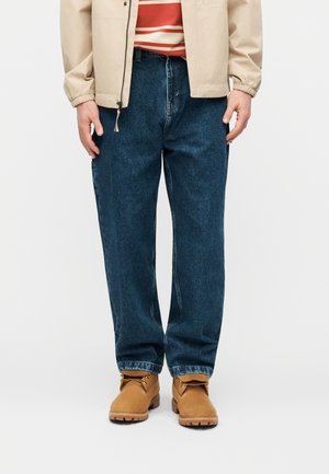 Mann trägt blaue Jeans mit geradem Bein, braune Arbeitsstiefel, beige Jacke und rot-weiß gestreiftes Hemd, steht vor einem weißen Hintergrund.