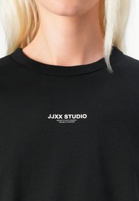 Black cotton t-shirt with a crew neck and short sleeves. Features white text logo "JJXX STUDIO" printed on the chest. Smooth texture.