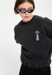 Mujer con gafas de sol negras y una sudadera negra desgastada con un pequeño bordado de un OVNI y un cactus en el pecho.