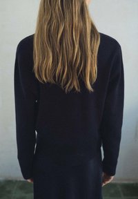 Čierny sveter vyrobený z mäkkého materiálu, s dlhými rukávmi a voľným strihom. Na chrbte sú dlhé, vlnité vlasy padajúce nadol.