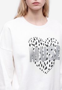 Jeune personne portant un sweat-shirt blanc avec texte argenté "MUSE" sur un cœur noir tacheté et les phrases "Rock and Love" et "Modern Nature."