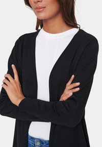 Črn kardigan z odprtim sprednjim delom, rebrasto teksturo, dolgimi rokavi, oblečenega čez belo majico in v kombinaciji z modrimi kavbojkami.