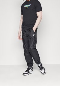 Svart t-shirt med flerfärgad logotyp, tillsammans med svarta glänsande byxor och svart-vita Nike-sneakers med elastiska muddarna.