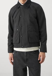 Sort knapjakke med klaplommer over en hvid skjorte, båret med sorte denimjeans på en stående person foran en ensfarvet baggrund.