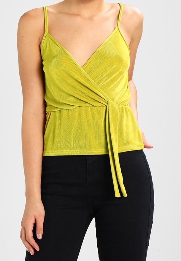 Femme portant un haut cache-cœur sans manches vert citron avec des bretelles fines et un pantalon noir, sur un fond clair uni.