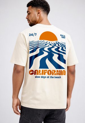 Man wearing off-white T-shirt with blue wavy pattern, orange sun, text "CALIFORNIA slow days at the beach," and "24/7 NVLS 1987."