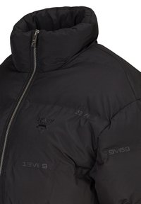 Zwarte pufferjas met een hoge kraag, voorzien van een zilveren rits en gebrandmerkte opdruk aan de voorkant. Glad, gewatteerd textuur overal.