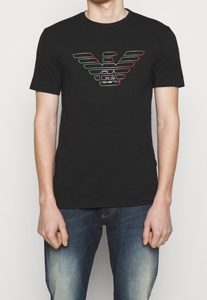 Mężczyzna w czarnej koszulce z stylizowanym logo orła w zielonym i czerwonym konturze oraz literami "GA", w połączeniu z niebieskimi, wyblakłymi jeansami.