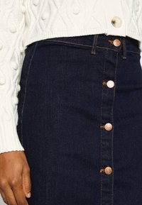 Mörkblå denimkjol med kopparfärgade knappar och kontrasterande sömmar, kombinerad med en krämfärgad stickad tröja med texturerade mönster.