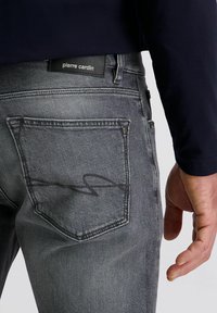 Vue arrière d'une personne portant un jean gris Pierre Cardin avec une étiquette de marque visible et une poche arrière, la main détendue sur le côté, en haut bleu marine.