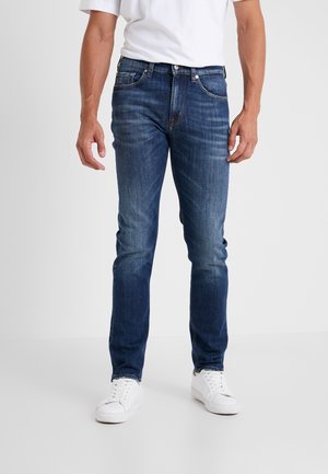 Homme portant un jean slim bleu, des baskets blanches et un t-shirt blanc, debout devant un fond uni.