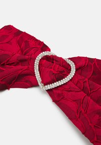 Sarkanas auduma lente ar tekstūru rožu dizainu, ar sudraba sirds formas sprādzi, kas rotāta ar caurspīdīgiem rhinestones.