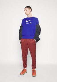 Plava Nike dukserica s logom "AIR", bordo jogger hlače i narančasta obuća s crnim akcentima. Outfit uključuje crnu jaknu.