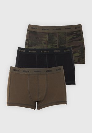 Stehender Mann in olivgrüner Boxershorts blickt nach links, posiert barfuß vor schlichtem weißem Hintergrund und zeigt die Passform der Unterwäsche.