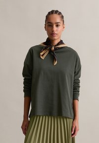 Donna con capelli intrecciati che indossa una felpa verde scuro, una gonna a pieghe oliva e una sciarpa decorata legata attorno al collo.