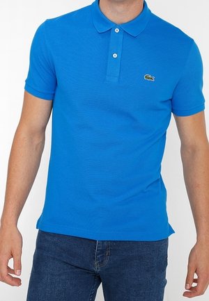 Homme portant un polo bleu vif à manches courtes avec un col et deux boutons, associé à un jean bleu foncé.