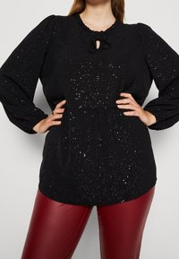 Haut noir à manches longues avec un col volanté, un détail à nouer et des paillettes dispersées. Associé à un pantalon en simili cuir rouge.