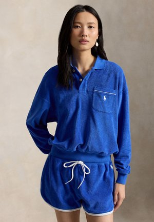 Ung kvinde med langt, mørkt hår iført en blå frotté langærmet polo og matchende shorts med hvid snor og kant.