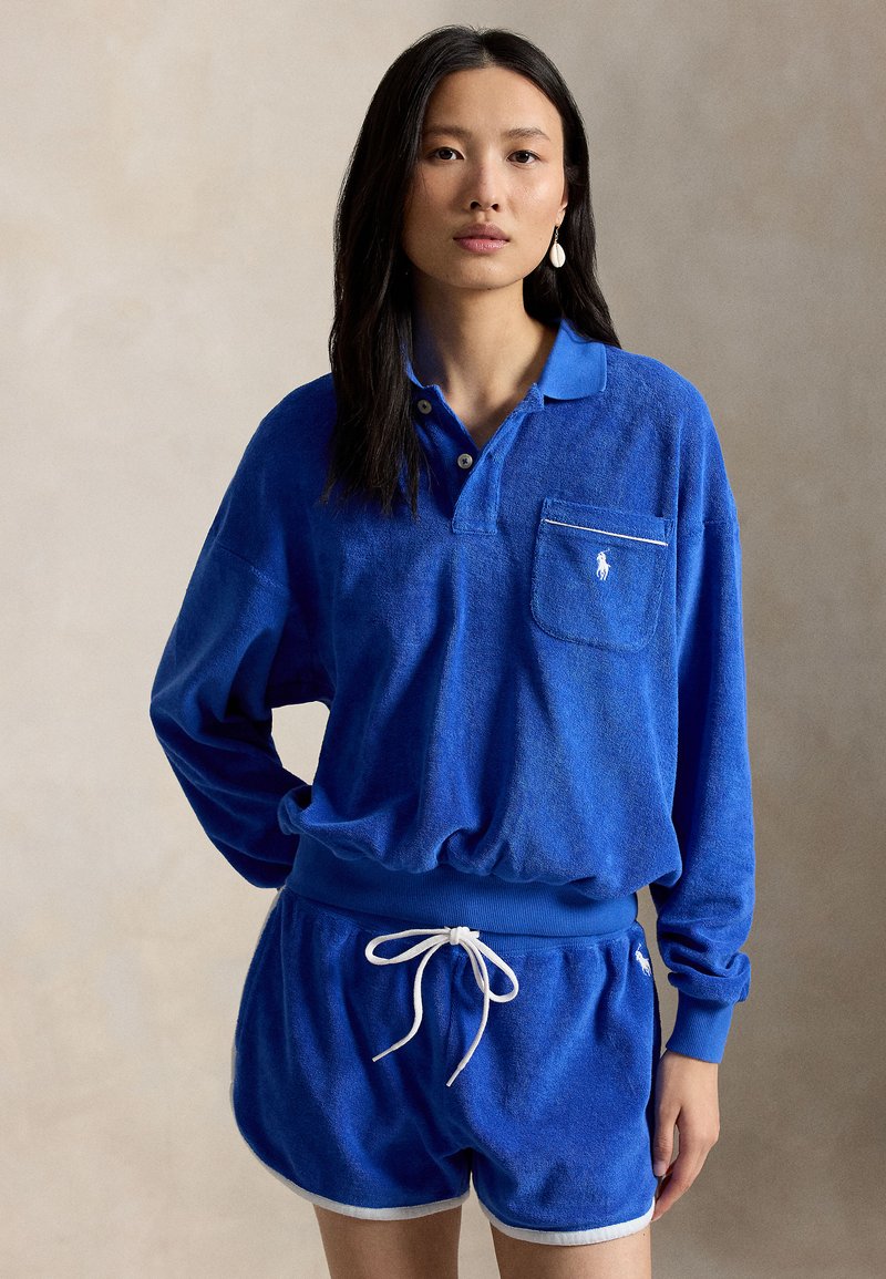 Mujer joven con cabello largo y oscuro que lleva un polo de manga larga de felpa azul y pantalones cortos a juego con cordón blanco y ribete.