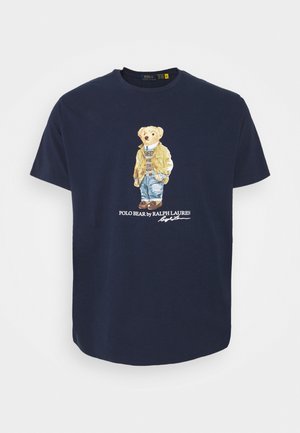 Marineblaues T-Shirt mit der Grafik eines Bären, der eine beige Jacke, einen Pullover, Jeans und Stiefel trägt, mit dem Text "POLO BEAR by RALPH LAUREN."