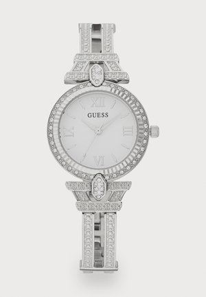 Srebrna analogna ura z belo številčnico, rimskimi številkami, okrašenim okvirjem in dekorativnim zapestnicom, ki vsebuje karlovnike in geometrijske poudarke.