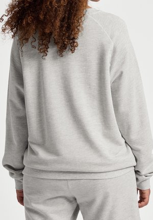 Femme aux cheveux bouclés portant un sweat-shirt gris clair et un pantalon de jogging assorti, vue de dos sur un fond uni.