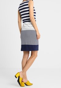 Raidallinen mekko, jossa on tummansinisiä, valkoisia ja harmaita vaakaraitoja. A-linjainen siluetti, ilman hihoja. Yhdistetty keltaisiin ja mustiin korkokenkiin.