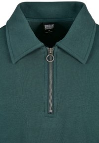 Zelena polo majica s patentnim zatvaračem sprijeda, širokim ovratnikom i teksturiranom tkaninom. Etiketa prikazuje "URBAN CLASSICS", veličina M, proizvedeno u Kini.