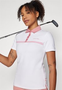Biely polo tričko s ružovým golierom a tromi gombíkmi. Obsahuje ružové horizontálne pruhy a logo Calvin Klein. Golfová palica držiaca na pleci.