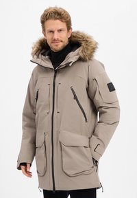 Beige parka met een nepbont kap, voorzien van meerdere rits- en klepzakken, een sluiting met rits aan de voorkant en slanke zwarte accenten. Zachte textuur.