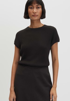 Vrouw met kort zwart haar die een zwart geribd kort mouwtopje en een zwarte rok draagt, staande tegen een effen lichte achtergrond.