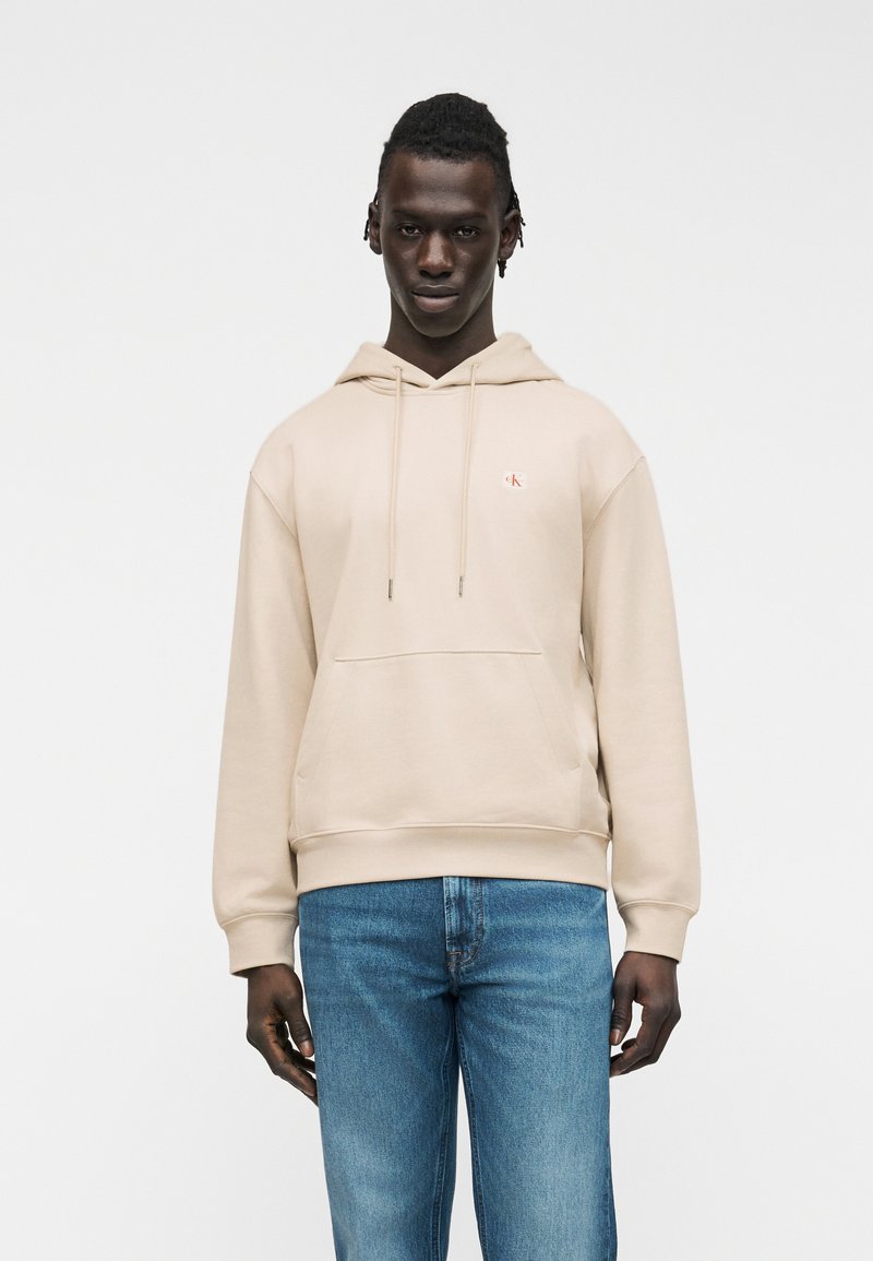 Άνδρας που φοράει μπεζ φούτερ με κουκούλα και μικρό λογότυπο στο στήθος, και μπλε τζιν, στέκεται μπροστά σε απλό ανοιχτό γκρι φόντο.