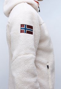 Sidläge på en person som bär en vit fleecjacka med norsk flagga som patch på överarmen och en dragkedjefickan nedanför.