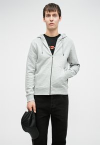 Hanorac gri deschis cu fermoar, cu glugă ajustabilă, buzunare frontale, purtat peste un tricou negru, asociat cu blugi negri și ținând în mână o șapcă neagră.