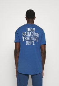 Blauwe katoenen T-shirt met een afgeronde zoom, met op de achterkant witte tekst: "IRON PARADISE TRAINING DEPT." in verschillende lettergroottes.