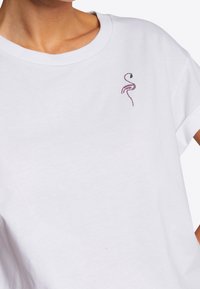T-shirt bianco in cotone con un piccolo design di un fenicottero in strass viola sul lato sinistro del petto. Maniche corte e vestibilità rilassata.
