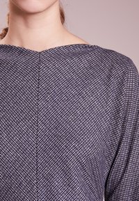 Haut bleu marine avec un motif tissé texturé, encolure bateau et coupe ample. Le tissu a un léger brillant et des détails de surpiqûre en diagonale.