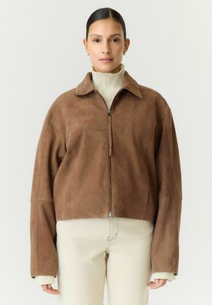 Frau trägt eine braune Wildlederjacke mit Reißverschluss und langen Ärmeln über einem weißen Rollkragenpullover und hellbeigen Hosen.