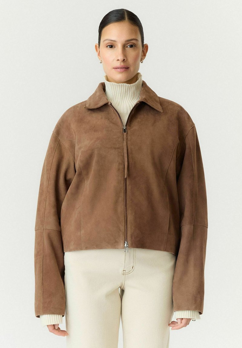 Femme portant une veste zippée en daim marron à manches longues par-dessus un pull à col roulé blanc et un pantalon beige clair.