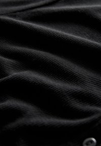 Black ribbed fabric featuring subtle texture variations, with a glimpse of metallic button detail, creating a soft, slightly stretchy appearance.